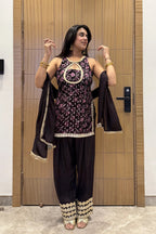 A woman stands indoors in front of a wooden door wearing a dark traditional outfit with a floral patterned top, loose dark pants, and a draped dupatta, accessorized with gold jewelry.