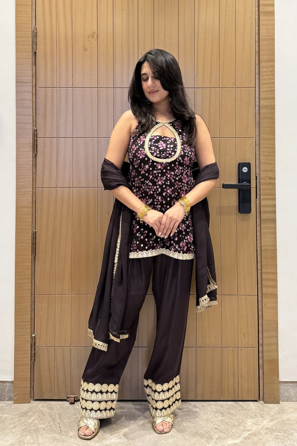 A woman stands in front of a wooden door wearing a dark brown ethnic outfit with a floral printed kurta, wide-leg trousers, and a draped dupatta, accessorized with gold jewelry.