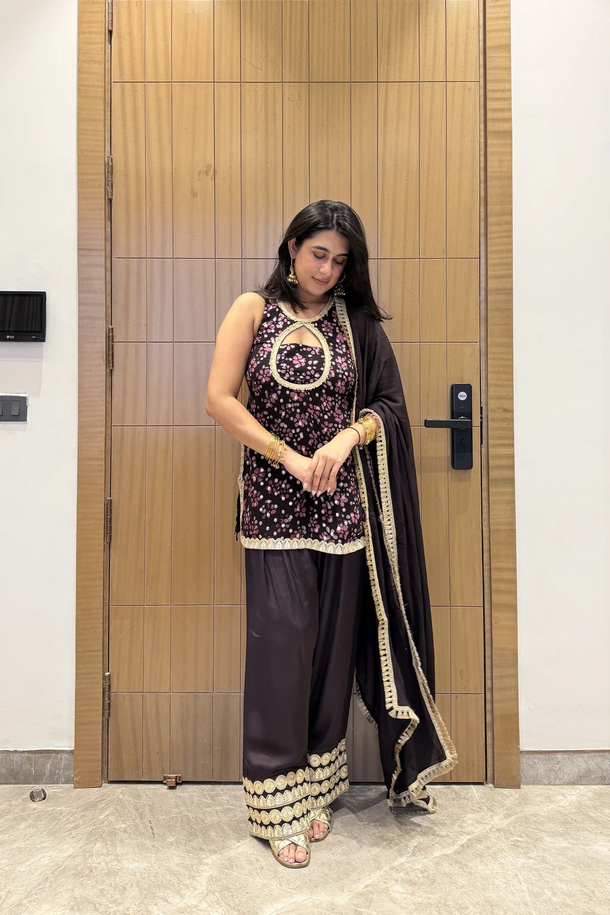 A woman modeling a traditional Indian outfit consisting of a black and purple floral printed kurta with gold embroidery, wide-leg dark purple palazzo pants with gold trim, and a black dupatta with gold borders, standing in front of a wooden door.