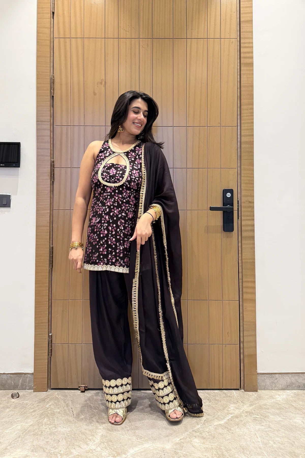 A woman models a traditional Indian outfit consisting of a black sleeveless tunic with pink floral embroidery and gold trim, black wide-leg pants with gold embroidered borders, and a black dupatta with gold trim, standing in front of a wooden door.